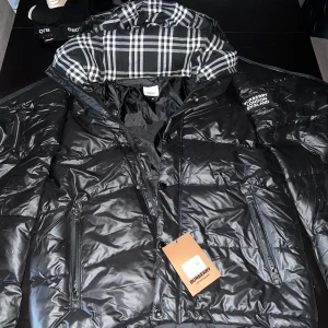 Svart pufferjacka från Burberry - Säljer en svart pufferjacka från Burberry i helt ny  skick. Jackan har en klassisk rutig insida i huvan och dragkedja framtill. Perfekt för kalla dagar och ger en stilren look. Den är inte äkta men det ganska bra kvalite på den. Storleken är M men passar mer till L eller XL.