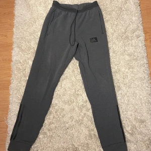 Grå mjukis/träningsbyxor från Adidas - Säljer ett par grå mjukisbyxor från Adidas i mycket bra skick. De har en bekväm passform med resår och snörning i midjan. Perfekta för träning eller chill hemma. Byxorna har en diskret Adidas-logga på benet och dragkedjor vid bensluten.