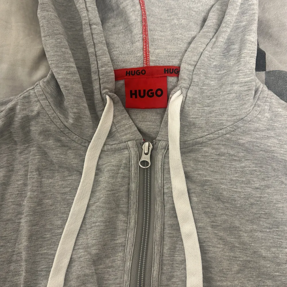 Säljer en snygg grå hoodie från Hugo i mycket bra skick. Den har en dragkedja framtill och vita snören i huvan. Perfekt för en avslappnad stil och passar till de flesta tillfällen. Stor logga på nederkanten ger en cool touch. Perfekt för höst och vår!. Tröjor & Koftor.