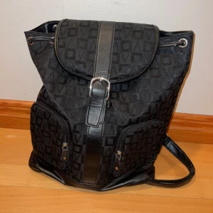 Svart ryggsäck med mönster - Säljer en snygg svart ryggsäck med diskret mönster och flera praktiska fack. Väskan har dragkedjor och en stilren design med ett spänne framtill. Perfekt för både vardag, utflykter och skolan! Från Don Donna. 