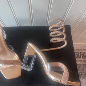 Guldiga sandaler med glittrig spiral - Snygga guldiga sandaler med en unik glittrig spiral som slingrar sig upp längs benet. De har en transparent klack och en glittrig rem över tårna. Perfekta för festliga tillfällen när du vill sticka ut och glänsa!