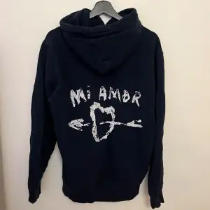 Säljer en mörkblå hoodie från Mira Paris med texten 'Mi Amor' och ett hjärta med pil på ryggen. Den har en klassisk känguruficka och justerbar huva med dragsko. Den har används några gånger men är fortfarande i bra skick. Köptes för 1200kr och är normal i passformen🤍 