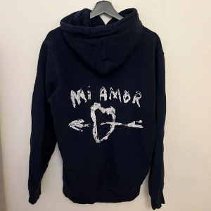 Mörkblå hoodie från Mira Paris - Säljer en mörkblå hoodie från Mira Paris med texten 'Mi Amor' och ett hjärta med pil på ryggen. Den har en klassisk känguruficka och justerbar huva med dragsko. Den har används några gånger men är fortfarande i bra skick. Köptes för 1200kr och är normal i passformen🤍 