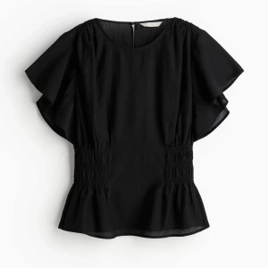 Svart blus - Snygg svart blus ifrån H&M som var jätte populär, finns inte längre på hm. Volangdetaljer i midjan och korta ärmar. Perfekt för både vardag och fest. Den har en smickrande passform och är superbekväm att bära. Använt 2 gånger så är bra skick och inga defekter.