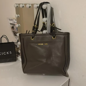 Brun skinnväska från Michael Kors - Säljer en snygg brun skinnväska från Michael Kors i mycket bra skick. Väskan har guldfärgade detaljer och flera praktiska fack inuti med ett stilrent mönster. Perfekt för både vardag och fest! 👜✨ej äkta