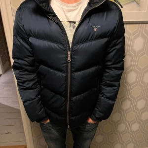 Mörkblå dunjacka från GANT - Säljer en snygg mörkblå dunjacka från GANT i storlek S. Jackan har en dragkedja framtill och är quiltad för extra värme. Perfekt för kalla höst- och vinterdagar. Den har en huva och är i mycket bra skick. Passar både till vardags och lite finare tillfällen.