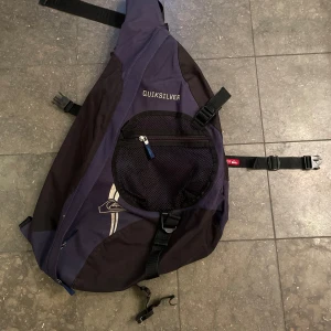 Svart och blå slingbag från Quiksilver - Säljer en cool svart och blå slingbag från Quiksilver. Perfekt för vardagsbruk eller när du är på språng. Väskan har en praktisk dragkedja och justerbara remmar för bästa passform. Den är i gott skick förutom ett spänne som är trasigt. 