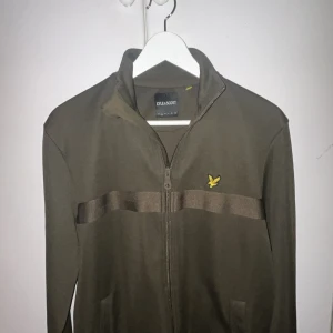 Olivgrön tröja från Lyle & Scott - Säljer en snygg olivgrön tröja från Lyle & Scott. Den har en dragkedja framtill och ett diskret märke på bröstet. Perfekt för höst och vår, med långa ärmar och en bekväm passform. Passar både till vardags och lite mer uppklädda tillfällen.