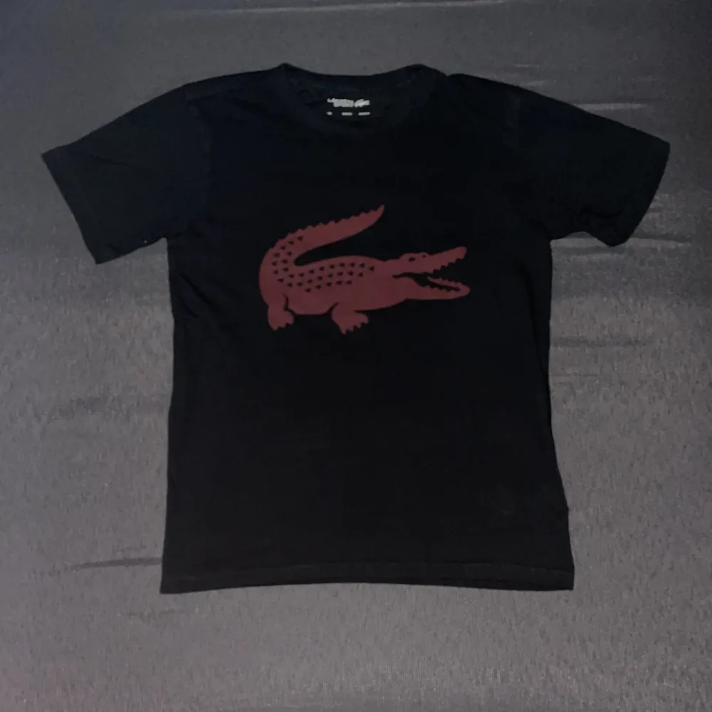 Säljer en svart t-shirt från Lacoste Sport med en stor röd krokodil på framsidan. Den är i bra skick och har korta ärmar. Materialet är Ultra Dry, perfekt för träning eller vardagsbruk. Passar både för sportiga aktiviteter och avslappnade dagar.. T-shirts.