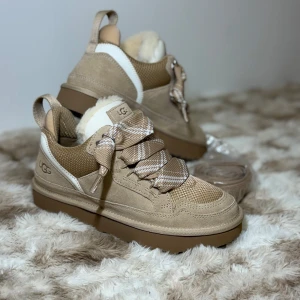 Beige sneakers från UGG - Säljer ett par snygga och bekväma beige UGG. De har en mysig pälsdetalj på insidan och breda snören med ett rutigt mönster. Perfekta för höst och vinter med sin robusta sula och varma insida. Skorna är i nytt skick.