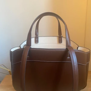 Brun handväska - Snygg handväska i brunt och vitt med stilren design. Väskan har två handtag och en praktisk mini necessär inuti för säker förvaring. Perfekt för både vardag och festliga tillfällen.