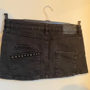 Svart jeanskjol med nitar - Snygg svart jeanskjol köpt secondhand med coola nitar på fickorna. Kjolen är kort och har en klassisk femficksdesign med knapp och dragkedja fram. Passar perfekt till både vardag och fest.