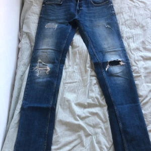 Blå jeans från Smog - Säljer ett par blå jeans från Smog i storlek 32/34. De har en cool slitning på knäna och en snygg tvättad look. Perfekta för en avslappnad stil. Passar bra till både vardag och fest. Dragkedja och knapp framtill.
