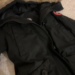 Svart parkajacka från Canada Goose - Säljer en svart parkajacka från Canada Goose i mycket bra skick. Jackan har en varm pälsfodrad huva och dragkedja framtill. Perfekt för kalla vinterdagar. Den är både snygg och funktionell, har några små dieffekter vid ärmarna därav priset. 