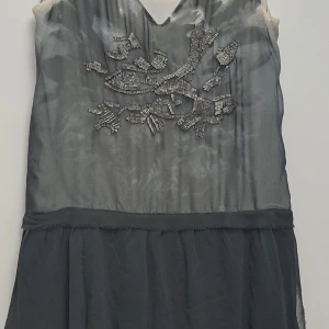 Grå klänning med pärldetaljer - Snygg grå klänning i 100% polyester med v-ringning och vackra pärldetaljer på bröstet. Perfekt för festliga tillfällen! Klänningen har en elegant design med en lätt transparent look och en mjuk känsla.