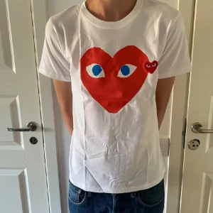 Vit t-shirt från Comme des Garçons - Säljer en vit t-shirt från Comme des Garçons med det ikoniska röda hjärtat med ögon på bröstet. T-shirten är i bomull och har en normal passform. Perfekt för en avslappnad stil eller att bära till vardags. Den är i mycket bra skick och redo för en ny ägare!