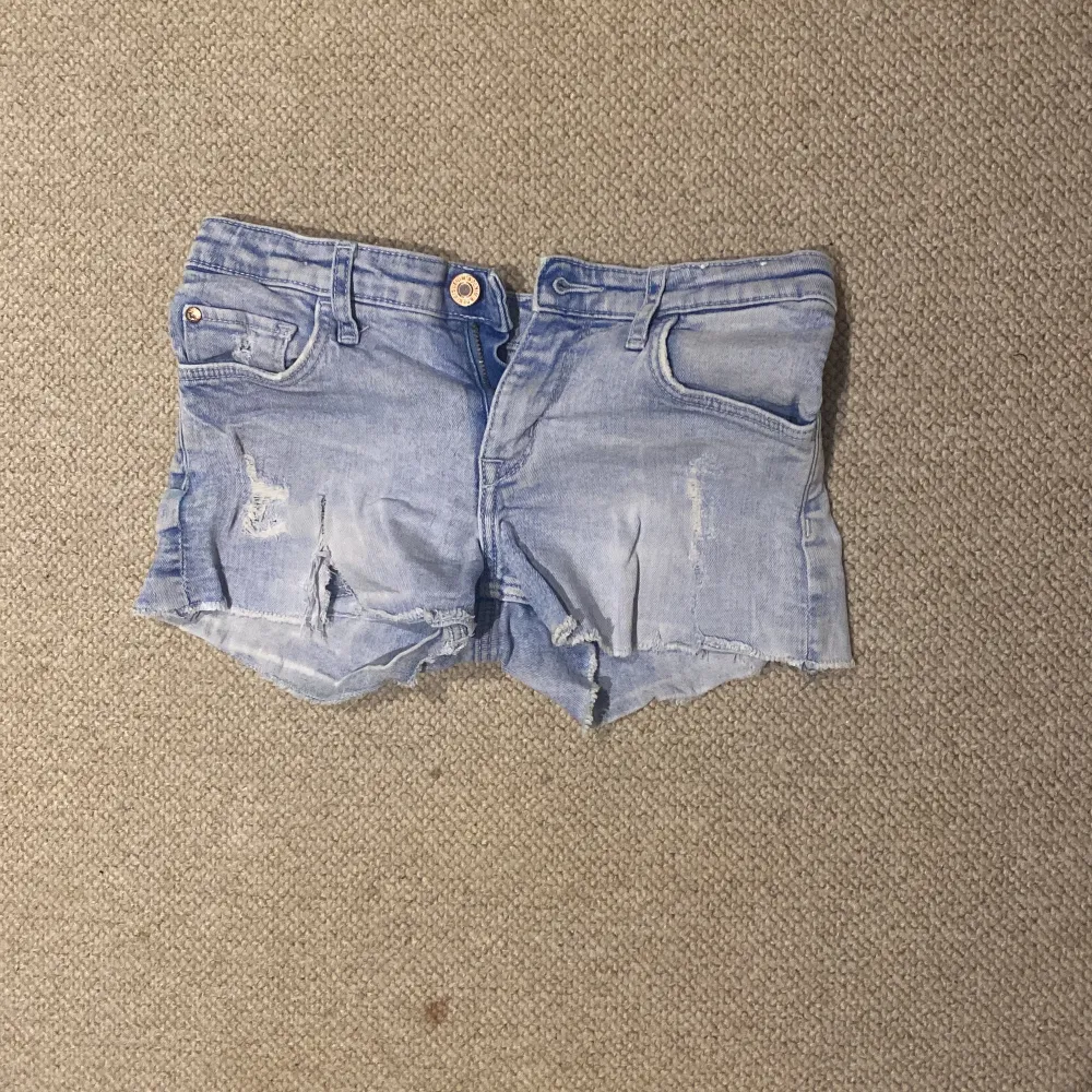 Säljer ett par ljusa jeansshorts i slitet skick med råa kanter och slitningar. Perfekta för sommardagar och festivaler! De har en knapp och dragkedja framtill samt klassiska fem fickor. Passar perfekt till en avslappnad stil.. Shorts.