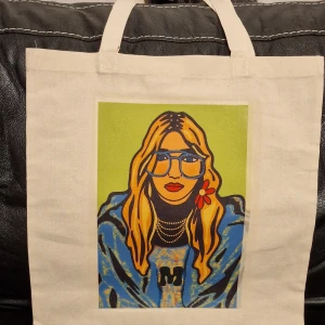 Tyg Beige shoppingväska med färgglatt tryck - Säljer en beige shoppingväska i bomull med ett coolt och färgglatt tryck av en person med glasögon och blommor. Perfekt för att bära med sig allt man behöver till skolan eller en dag på stan. Väskan har två handtag och är rymlig och praktisk.