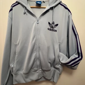 Ljusblå hoodie från Adidas - Säljer en snygg ljusblå hoodie från Adidas med dragkedja och klassiska tre ränder i mörkblått längs ärmarna. Den har en stor logga på bröstet och är perfekt för både träning och vardag. Superbekväm och i bra skick! Pris kan diskuteras!!💘