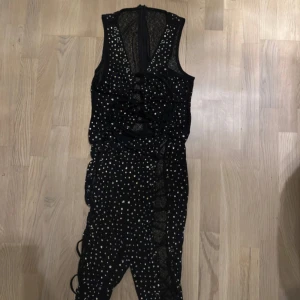 Svart glittrig jumpsuit med snörning - Snygg svart jumpsuit med glittriga detaljer och snörning längs benen. Perfekt för festliga tillfällen! Den har en tight passform och är gjord i ett syntetmaterial som ger en bekväm känsla. Denna jumpsuit är ett måste för dig som vill sticka ut på festen!