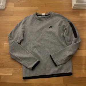 Grå Nike tech tröja - Säljer en snygg grå Nike tech i mycket bra skick (äkta). Den har en stilren design med svarta detaljer vid ärmslut och nederkant. Perfekt för både träning och vardag. Långärmad och gjord i ett mjukt material som känns skönt mot huden.