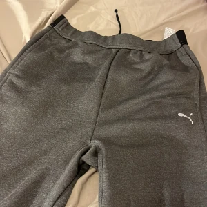 Grå mjukisbyxor från Puma - Säljer ett par grå mjukisbyxor från Puma i mycket bra skick. Byxorna har en bekväm passform med elastisk midja och svarta detaljer på sidorna. Perfekta för en avslappnad dag hemma eller lätt träning. 🐾