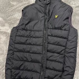 Svart dunväst från Lyle & Scott - Säljer en svart dunväst från Lyle & Scott i storlek 15/16 år. Passar även 13-14. Västen har en dragkedja framtill och är perfekt för höst och vår. Den är i mycket bra skick och har en liten gul logga på bröstet. Perfekt för att hålla sig varm och stilren!