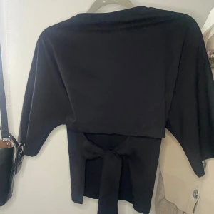 Svart topp från Nelly - Säljer en stilren svart topp från Nelly i storlek M. Toppen har en snygg knytning i ryggen som ger en extra detalj. Perfekt för både vardag och fest! 
