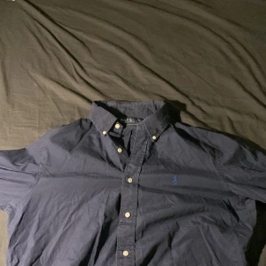 Mörkblå skjorta från Ralph Lauren - Säljer en snygg mörkblå skjorta från Ralph Lauren i storlek L. Skjortan har en klassisk krage och knappar fram. Den är broderad med den ikoniska Ralph Lauren-loggan på bröstet. Perfekt för både vardag och fest! 💙