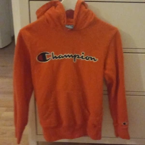 Orange hoodie från Champion - Säljer en cool orange hoodie från Champion i storlek XS. Den har en stor Champion-logga på bröstet och en praktisk magficka. Perfekt för höst och vår, eller när du bara vill chilla hemma. Huvan är skön och ger extra mysfaktor. Passar både till vardags och träning!