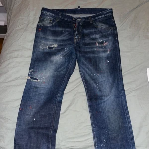 Blå jeans från DSQUARED2 - Säljer ett par coola blå jeans från DSQUARED2 med slitna detaljer och färgstänk för en edgy look. De har en knappgylf och klassisk femficksdesign. Perfekta för en avslappnad stil med lite attityd. Passar bra till både vardag och fest!