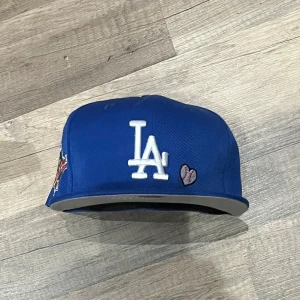 Blå keps från New Era - Säljer en blå New Era keps med broderad LA-logga och detaljer som '1988 World Series' och 'Love & Hustle = Game'. Kepsen är i polyester och har en cool stil med hjärtan och andra broderier. Perfekt för baseballfans eller som en snygg accessoar.