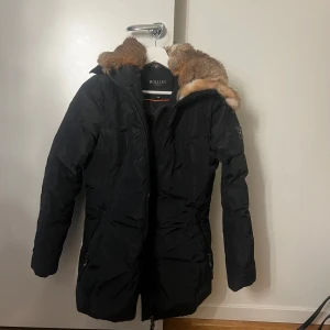 Svart vinterjacka med pälsdetaljer från Hollies - Säljer en snygg svart vinterjacka från Hollies i mycket bra skick, som ny. Jackan har en mysig pälsdetalj runt huvan och är perfekt för kalla vinterdagar. Den har en dragkedja framtill och långa ärmar. Supervarm och stilren! Den är använd ett få par gånger. Pris kan diskuteras!