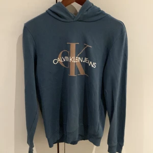 Blå hoodie från Calvin Klein Jeans - Säljer en snygg blå hoodie från Calvin Klein Jeans. Den har en stor logga på framsidan och är i mycket bra skick. Perfekt för höst och vår, med långa ärmar och en skön luva. Passar både till vardags och chillkvällar. 
