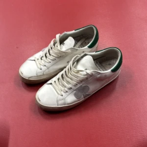 Vita sneakers med gröna detaljer - Säljer ett par vita sneakers i skinn med gröna detaljer och ett spader-märke på sidan. Skorna har snörning och är i bra skick, perfekta för vardagsbruk. Innersulan har ett inspirerande citat och färgglada stänk. Passar bra till både jeans och shorts!