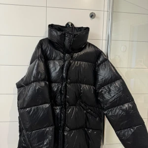 Svart pufferjacka från Peak performance  - Säljer en svart pufferjacka från Peak performance i mycket bra skick. Den är perfekt för kalla vinterdagar med sin tjocka vaddering och höga krage. Jackan har en dragkedja, både fram och vid sidorna, och knappar framtill för extra skydd mot kylan. Den är långärmad och har en glansig finish som ger en modern look. Perfekt för både vardag och vinteräventyr!