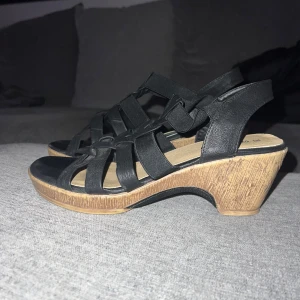 Svarta klackar med låg klack - Snygga svarta klackar med flätade remmar och en bekväm klack i trälook. Perfekta för sommarens alla tillfällen, både till vardags och fest. De har en öppen tå och elastiska remmar för en bra passform. Använd dem för att ge din outfit en stilren touch!