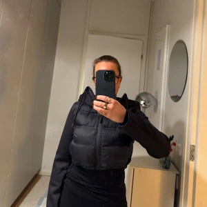 Svart kort pufferjacka med huva - Säljer en svart pufferjacka från Zara i storlek XS. Jackan har en praktisk huva och dragkedja framtill. Perfekt för kyliga dagar och ger en stilren look. Använd ett fåtal gånger och i mycket bra skick.