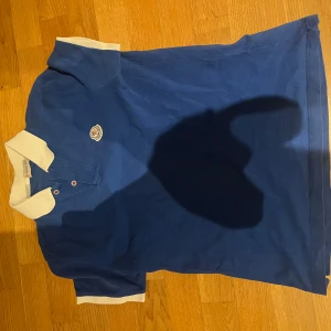Blå pikétröja från Moncler - Säljer en snygg blå pikétröja från Moncler i bra skick. Den har vita detaljer på kragen och ärmsluten samt en broderad Moncler-logga på bröstet. Perfekt för en stilren och avslappnad look. Passar både till vardags och festliga tillfällen.                                                                               Säljs inte längre!! Köpt på monclers hemsida för 2295kr. Passar 165-177 cm 