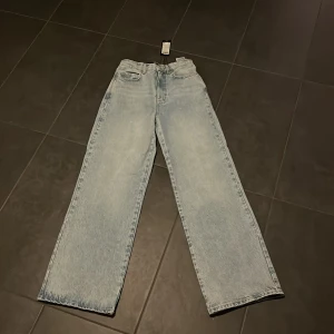 Ljusblå jeans från Vero Moda - Säljer ett par ljusblå jeans från Vero Moda i modellen 'Rebecca'. De är i nytt skick och har en snygg wide fit som är perfekt för både vardag och fest. Jeansen är tillverkade i denim och har en klassisk design med fem fickor och knappgylf. Perfekta för att styla med en t-shirt eller en mysig tröja!