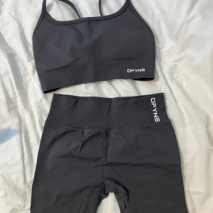 Grått träningsset från DFYNE - Snyggt träningsset från DFYNE med en sport-bh och matchande shorts. Bh:n har smala axelband och korsad rygg för extra stöd. Shortsen har hög midja och en rynkad detalj bak för en smickrande passform. Perfekt för gymmet eller yoga!