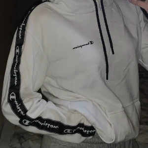 Vit hoodie från Champion - Säljer en vit hoodie från Champion. Den har svarta detaljer med Champion-loggan längs ärmarna och en broderad logga på bröstet.