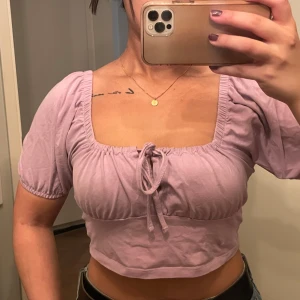 Ljuslila croppad topp - Säljer en superfin ljuslila croppad topp med korta puffärmar och en söt knytdetalj framtill. Perfekt för sommardagar eller en avslappnad fest. Toppen är i ett mjukt och bekvämt material som sitter skönt på kroppen.