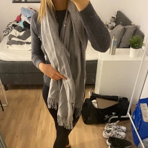 Grå halsduk med fransar - Säljer en superfin grå halsduk med fransar i ändarna. Den är perfekt för att hålla sig varm under höst och vinter. Halsduken är i ett mjukt material som känns skönt mot huden. Passar till de flesta outfits och är ett måste i garderoben! 🧣