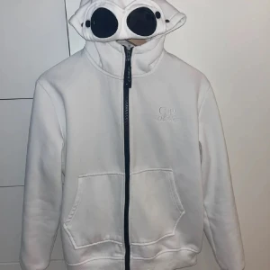 Vit hoodie med glasögon från C.P. Company - Säljer en unik vit hoodie från C.P. Company med inbyggda glasögon i huvan. Perfekt för dig som vill sticka ut! Den har en dragkedja framtill och praktiska fickor. Supercool och bekväm för både att gå ut i äta eller en vanlig dag i skolan