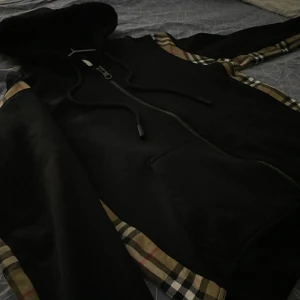 Burberry Graham Checked Panelled Zip-Up Hoodie - Säljer en snygg svart hoodie från Burberry med klassiska rutiga detaljer på ärmarna och är i mycket bra skick. Perfekt för höst och vinter väldigt tjock och varm. Den är storlek S men sitter typ som M så vilket som går lika bra.