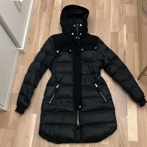 Svart dunjacka från J.Lindeberg - Säljer en snygg svart dunjacka från J.Lindeberg i storlek 34. Jackan har en hög krage och huva för extra värme. Den har långa ärmar och praktiska dragkedjor på fickorna. Perfekt för kalla vinterdagar! 🖤