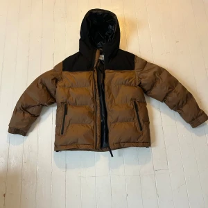 Brun och svart vinterjacka från Carhartt - Säljer en snygg brun och svart pufferjacka från Carhartt i storlek M. Jackan har en huva och dragkedja framtill, perfekt för kalla dagar. Den är i  bra skick och passar både höst och vinter. 🍂❄️