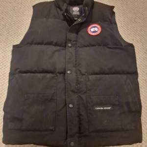 Säljer en svart dunväst från Canada Goose i bra skick. Västen har en klassisk design med knappar och en broderad logga på bröstet. Perfekt för kyliga höst- och vinterdagar.  Storlek L