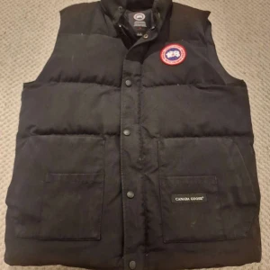 Svart dunväst från Canada Goose - Säljer en svart dunväst från Canada Goose i bra skick. Västen har en klassisk design med knappar och en broderad logga på bröstet. Perfekt för kyliga höst- och vinterdagar.  Storlek L